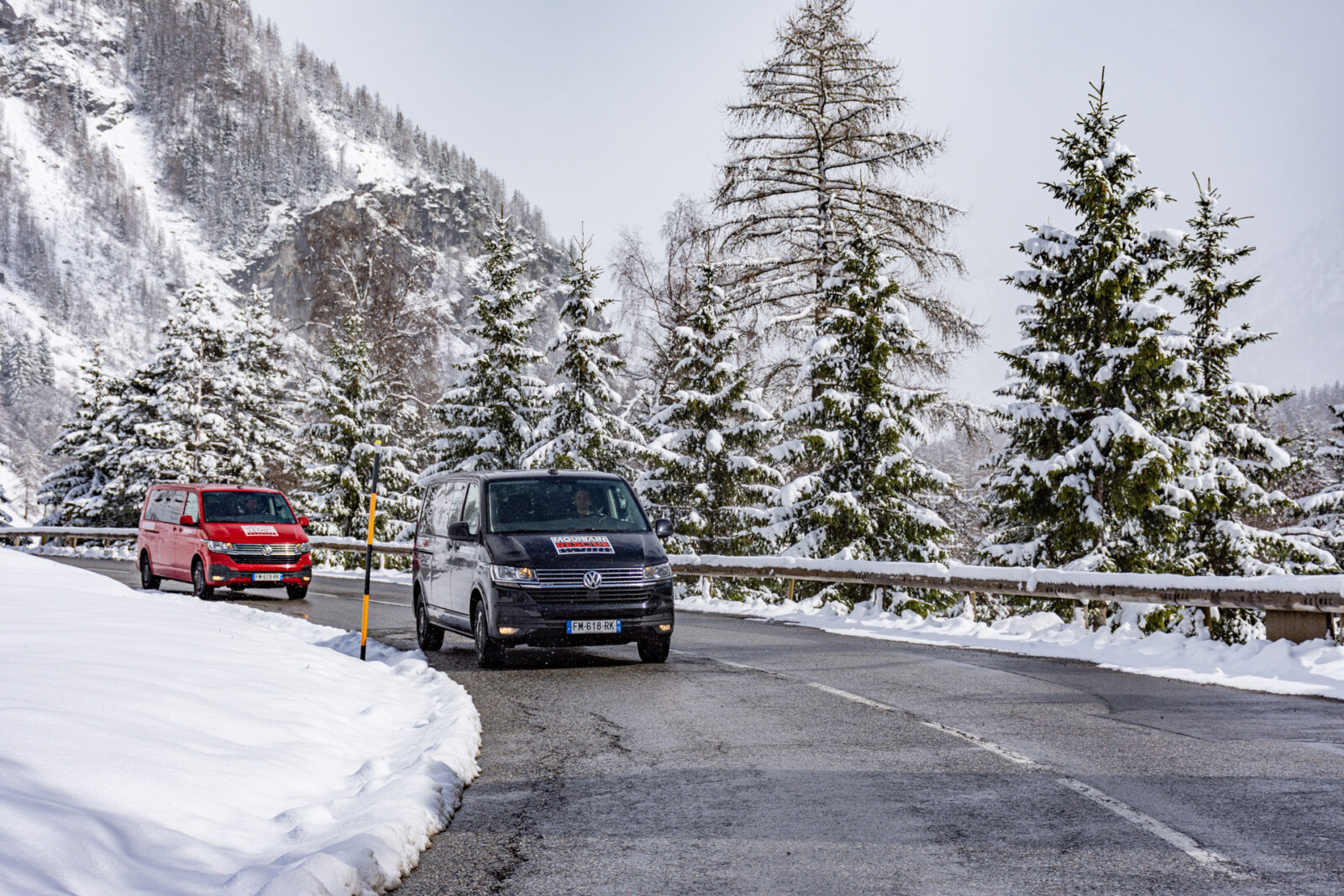 Red and black transfer vans driving on road surrounded by snow covered trees.