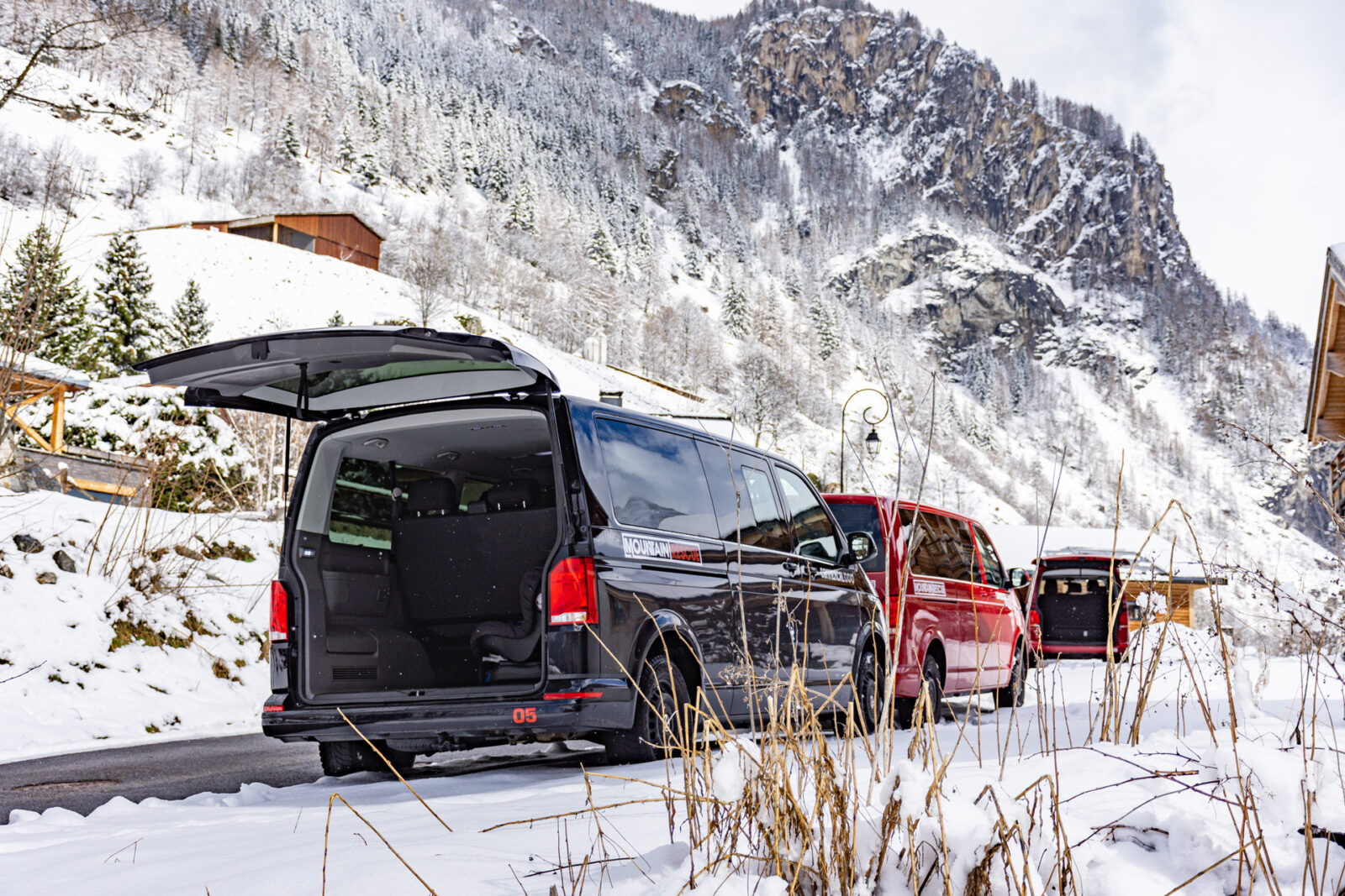 3 transfer vans with open boots parked on snow hill next to ski chalets