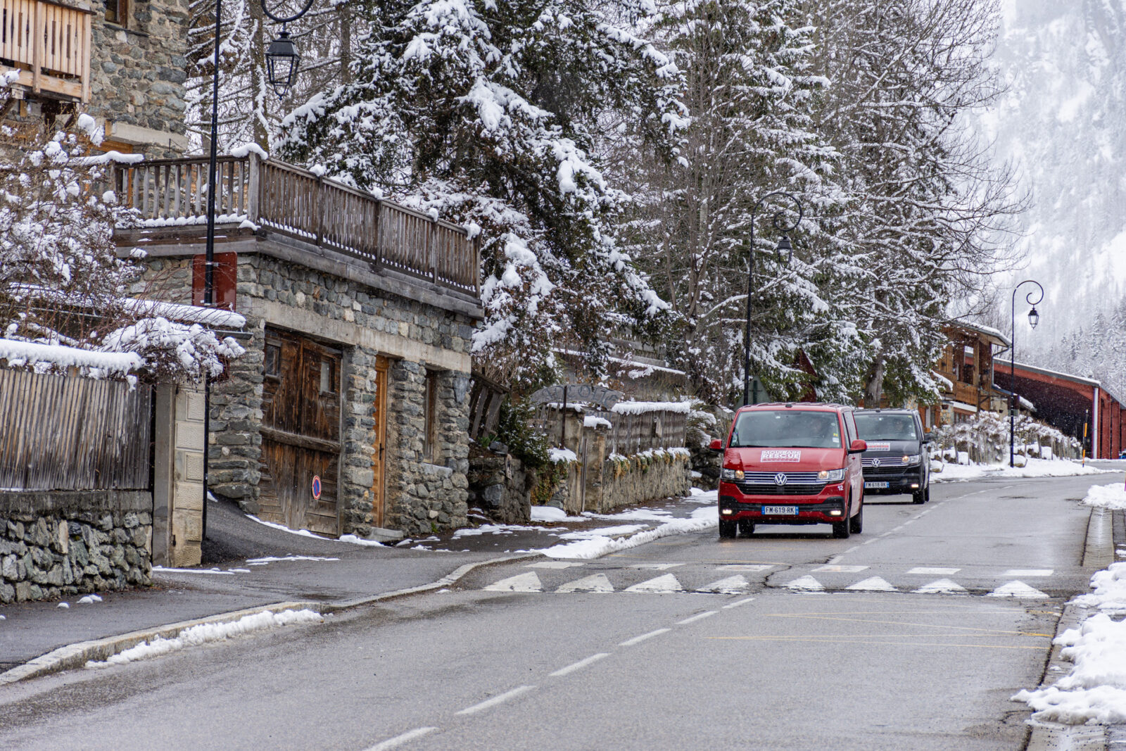 Two VW Vans driving down mountain village road in the winter